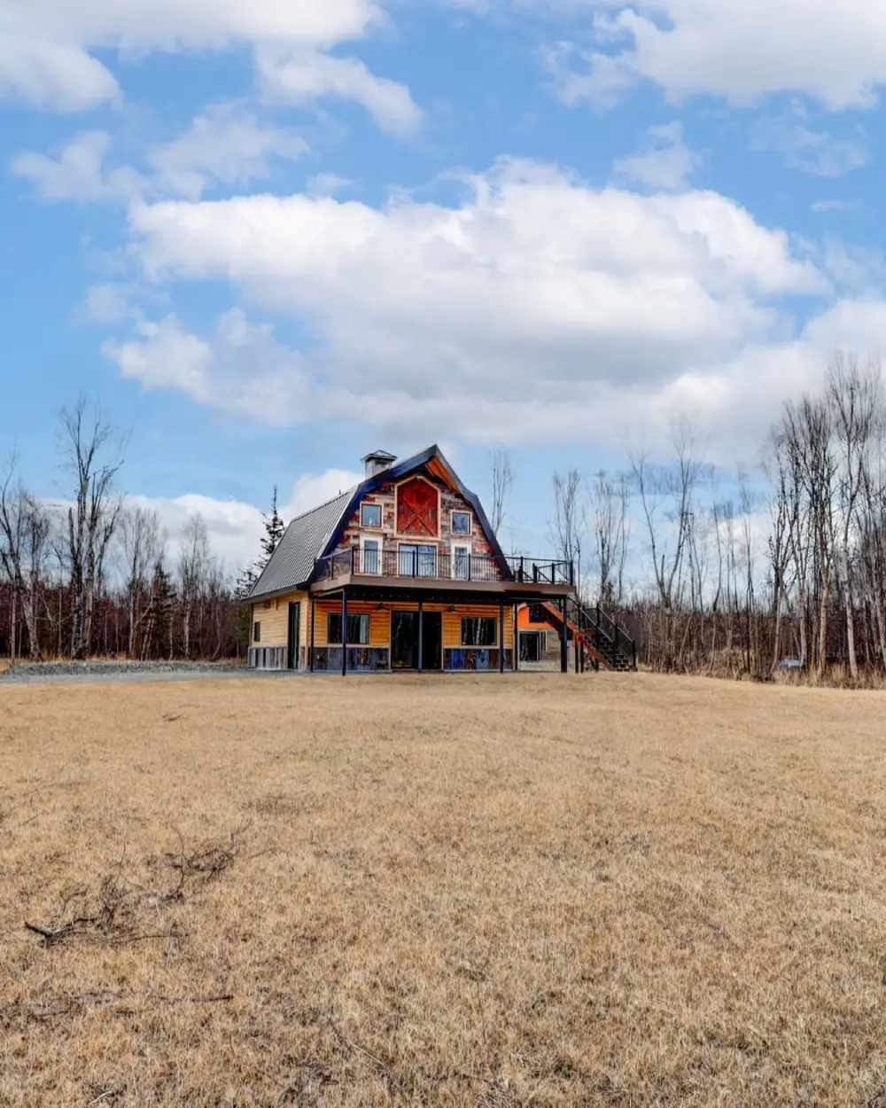Barn-style building with a steep metal roof and large windows sits in an open field, with leafless trees and a cloudy sky behind it. Borealis Bison Barns | Palmer, AK