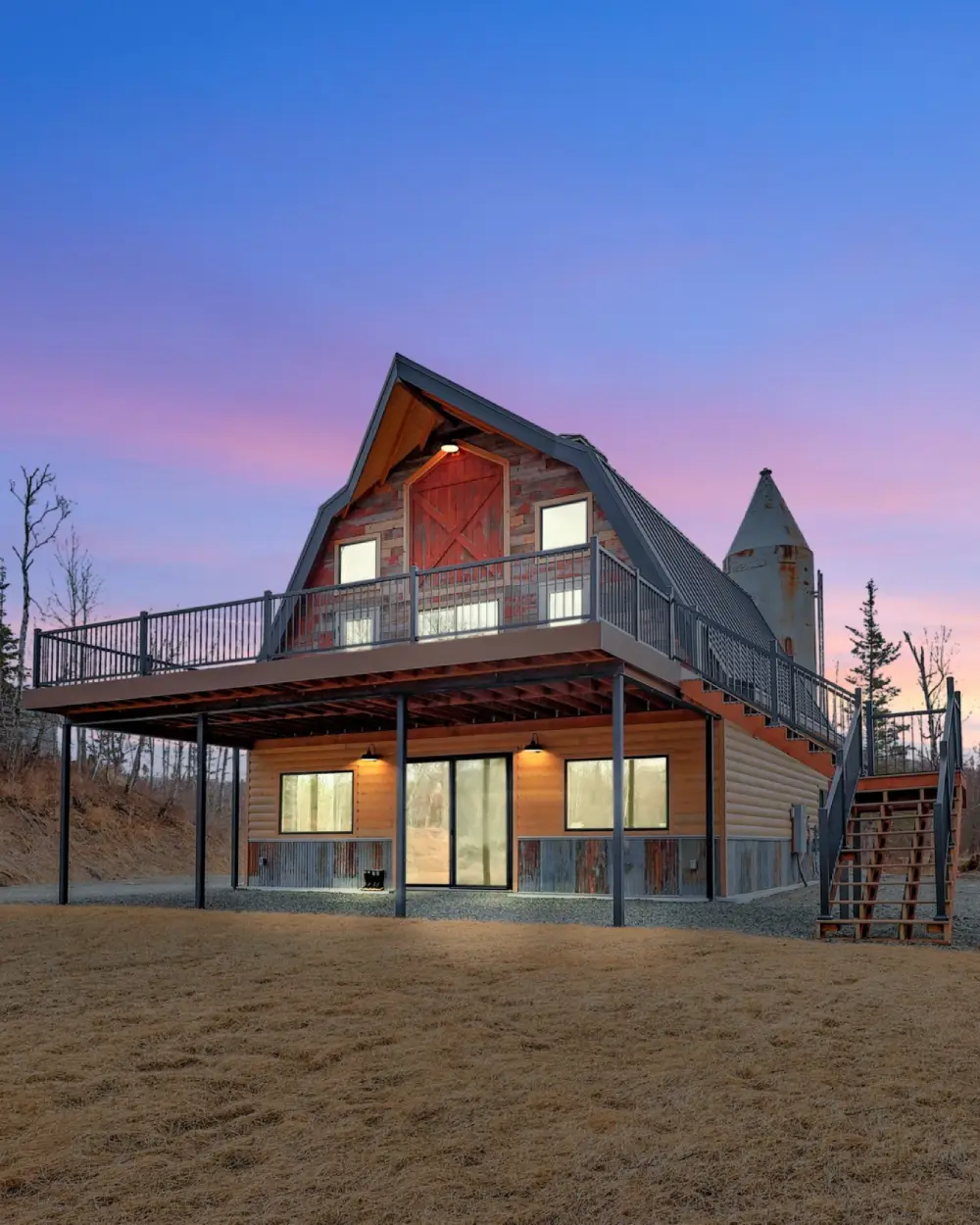 Barn-style building with elevated deck, exterior stairs, and silo, lit from inside at dusk under a pink and blue sky. Borealis Bison Barns | Palmer, AK