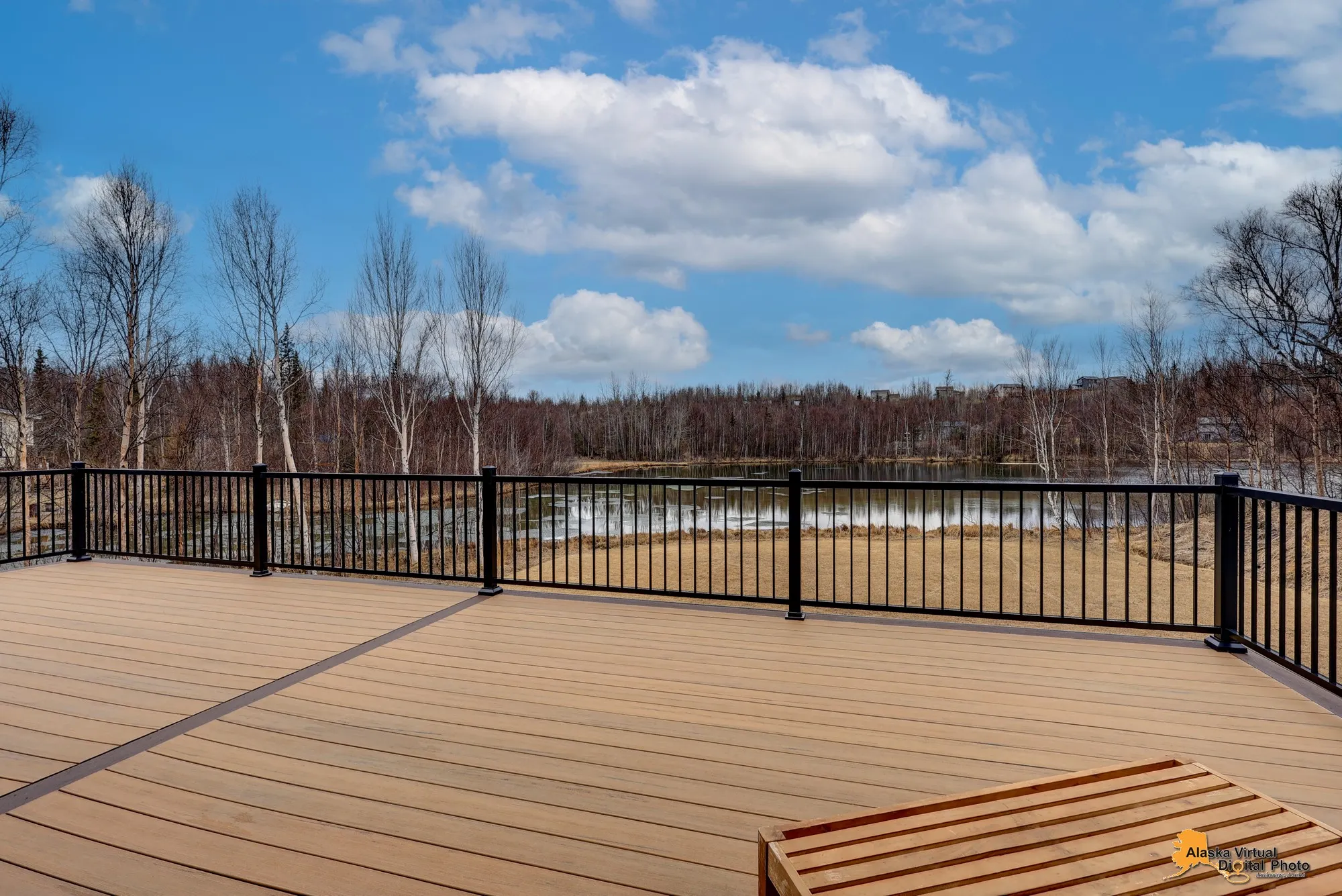 View from a wooden deck with black railing overlooking a pond and trees under a partly cloudy sky. Borealis Bison Barns | Palmer, AK
