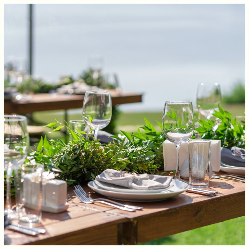 Outdoor table set with plates, glassware, and a green garland centerpiece, with another table blurred in the background. Borealis Bison Barns | Palmer, AK