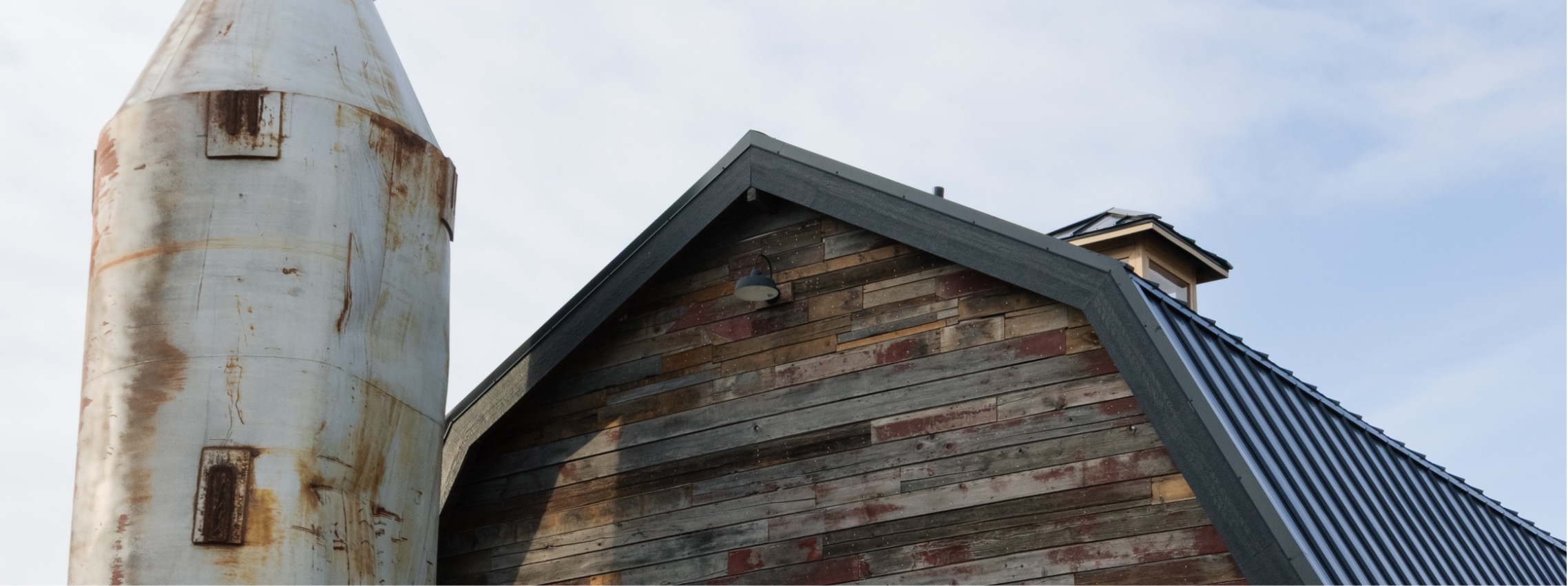 Close view of a rusted metal silo beside a barn-style building with weathered wood siding and metal roof under a cloudy sky. Borealis Bison Barns | Palmer, AK