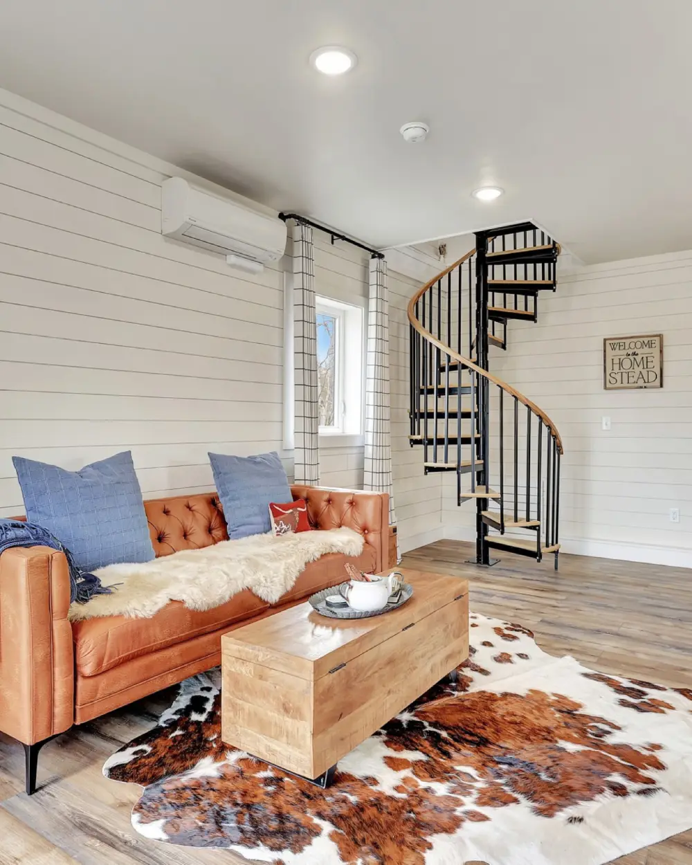 Cozy living area with orange sofa, wood floors, and spiral staircase, accented by a cowhide rug and neutral decor. Borealis Bison Barns | Palmer, AK