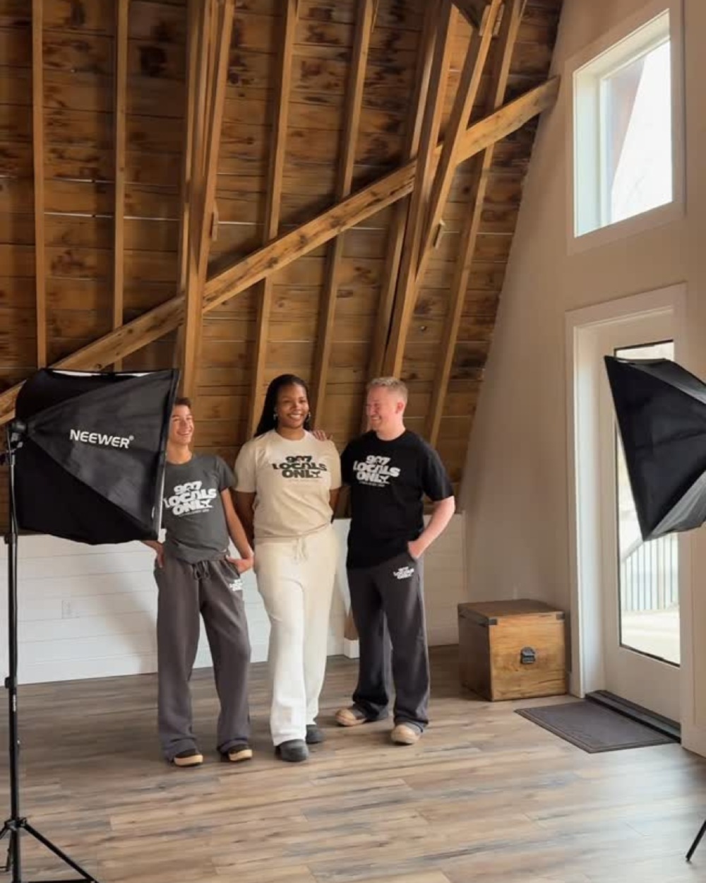 Three people stand in a barn-style interior with exposed wood beams and studio lights, wearing matching logo T-shirts. Borealis Bison Barns | Palmer, AK