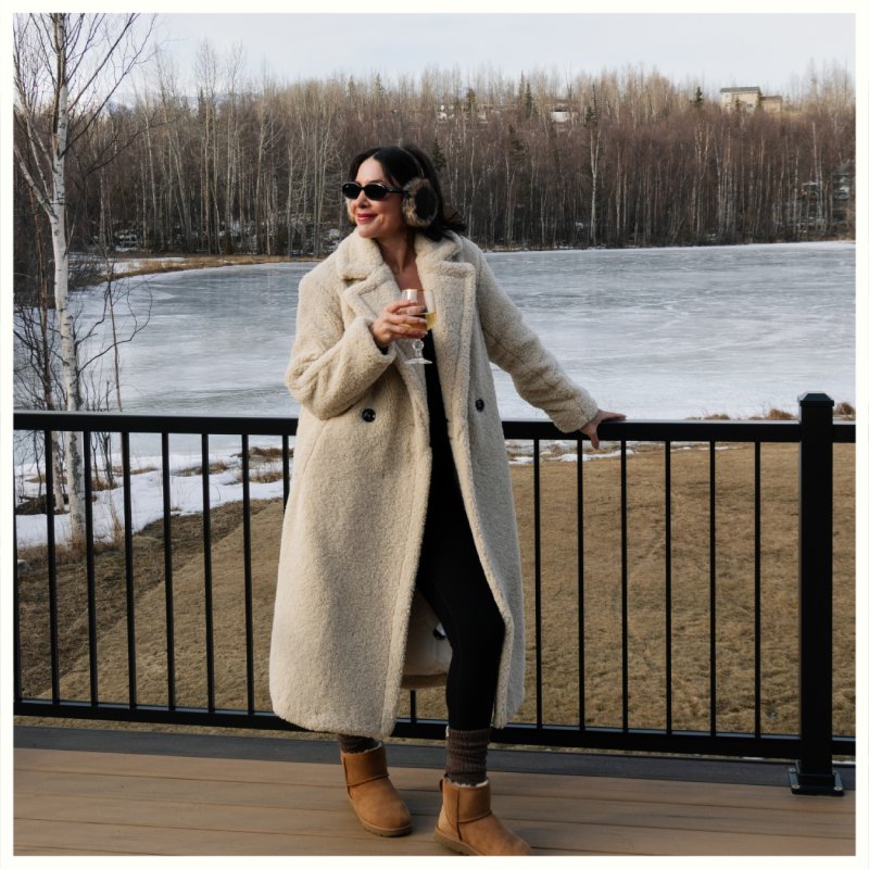 Person in a long coat and earmuffs stands on a deck holding a drink, with a partially frozen pond and trees behind. Borealis Bison Barns | Palmer, AK