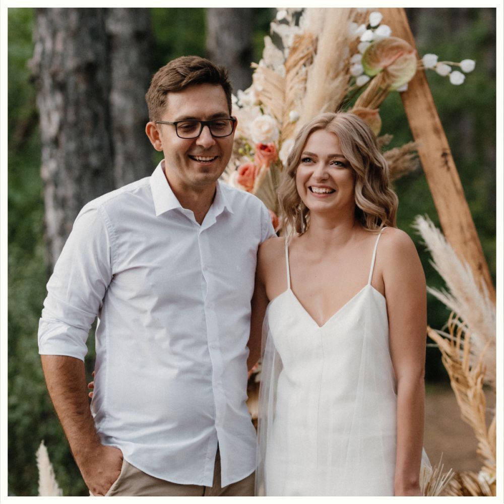 Two people stand smiling outdoors in front of a floral arch, one wearing a white dress and the other a white shirt. Borealis Bison Barns | Palmer, AK