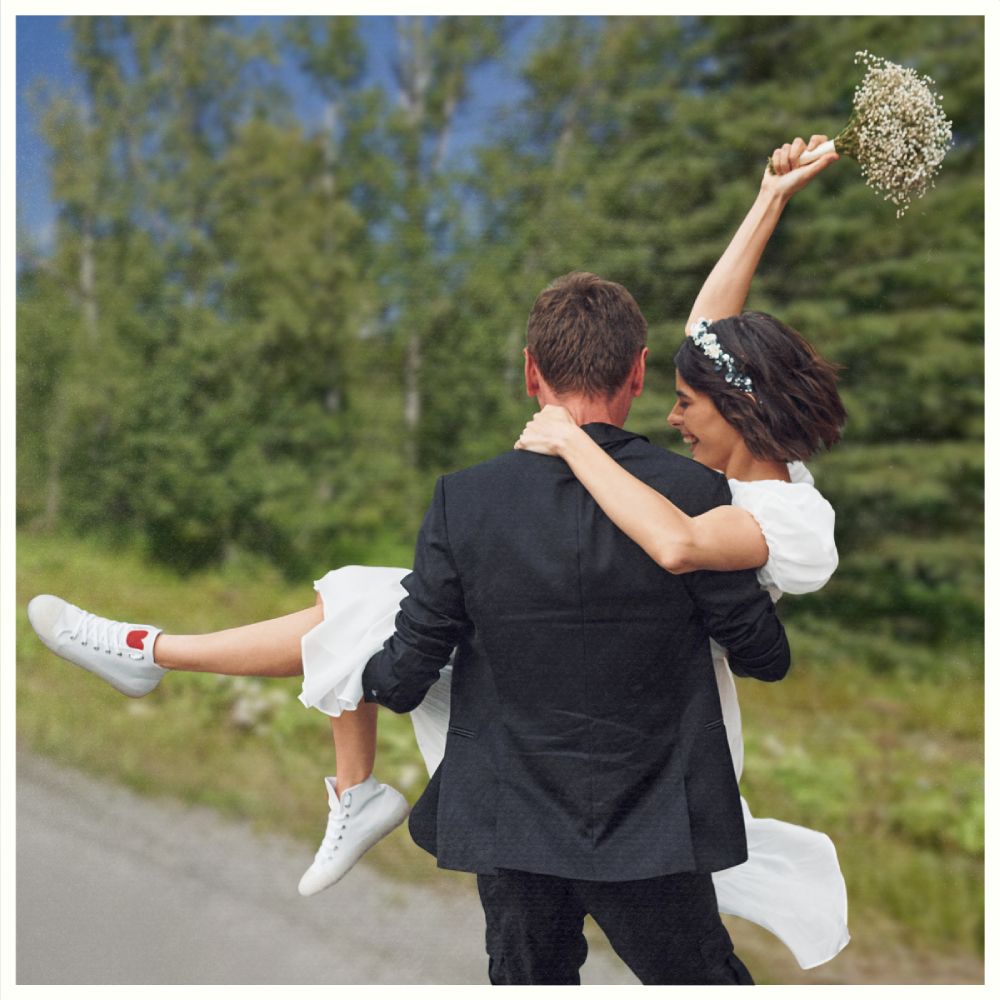 Person in a suit carries another person in a white dress holding flowers, outdoors on a road with blurred greenery behind. Borealis Bison Barns | Palmer, AK