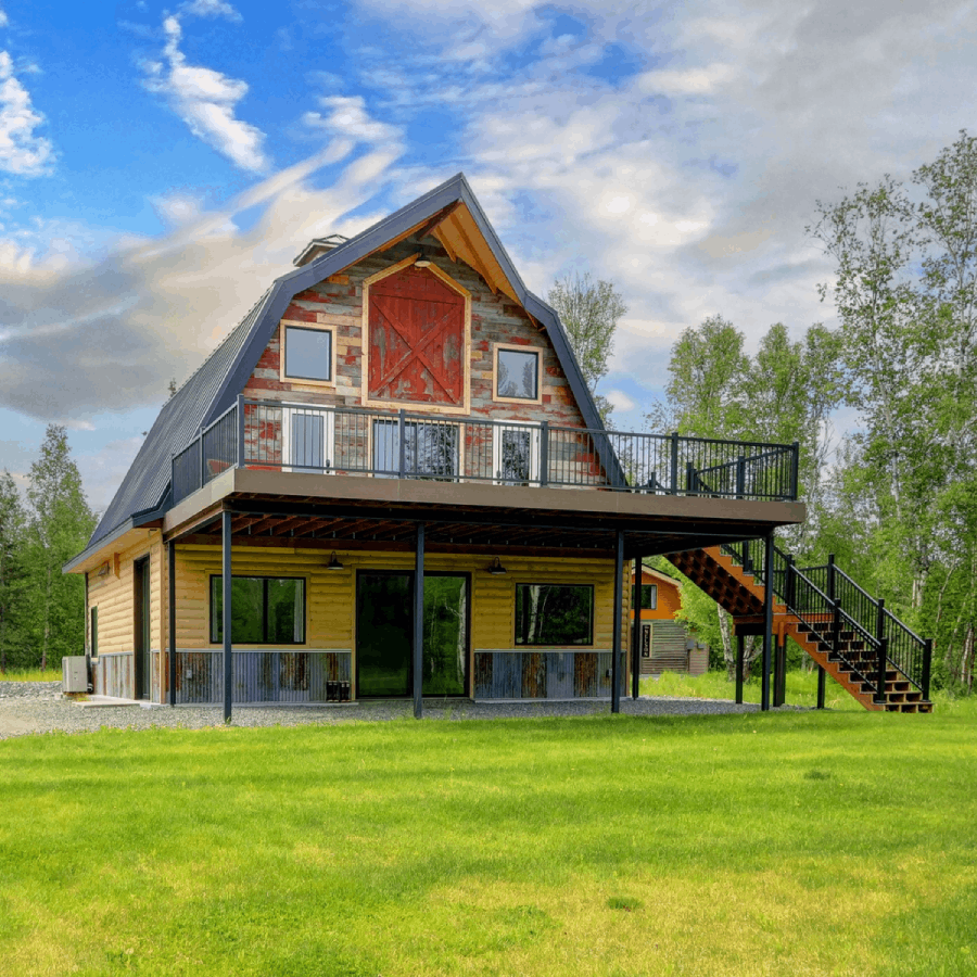 Exterior of the restored barn at Borealis Bison Barns in Alaska featuring a wraparound deck, rustic wood siding, and spacious lawn surrounded by trees.