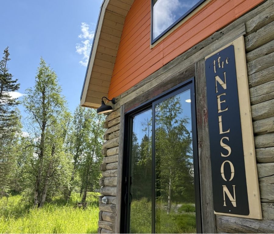 Close-up of The Nelson cabin entrance with log walls, black-framed glass doors, outdoor light, and vertical Nelson sign.
